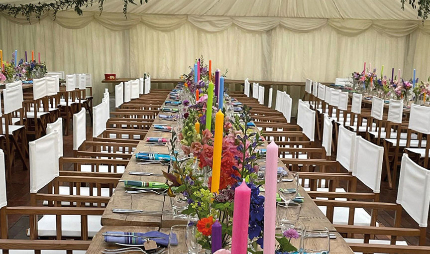 a long table in a wedding marquee with multicoloured flowers and candles and wooden director chairs