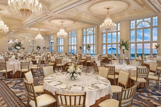 A grand ballroom with chandeliers, round tables with white tableclothes, white chairs and white flowers as a centrepiece