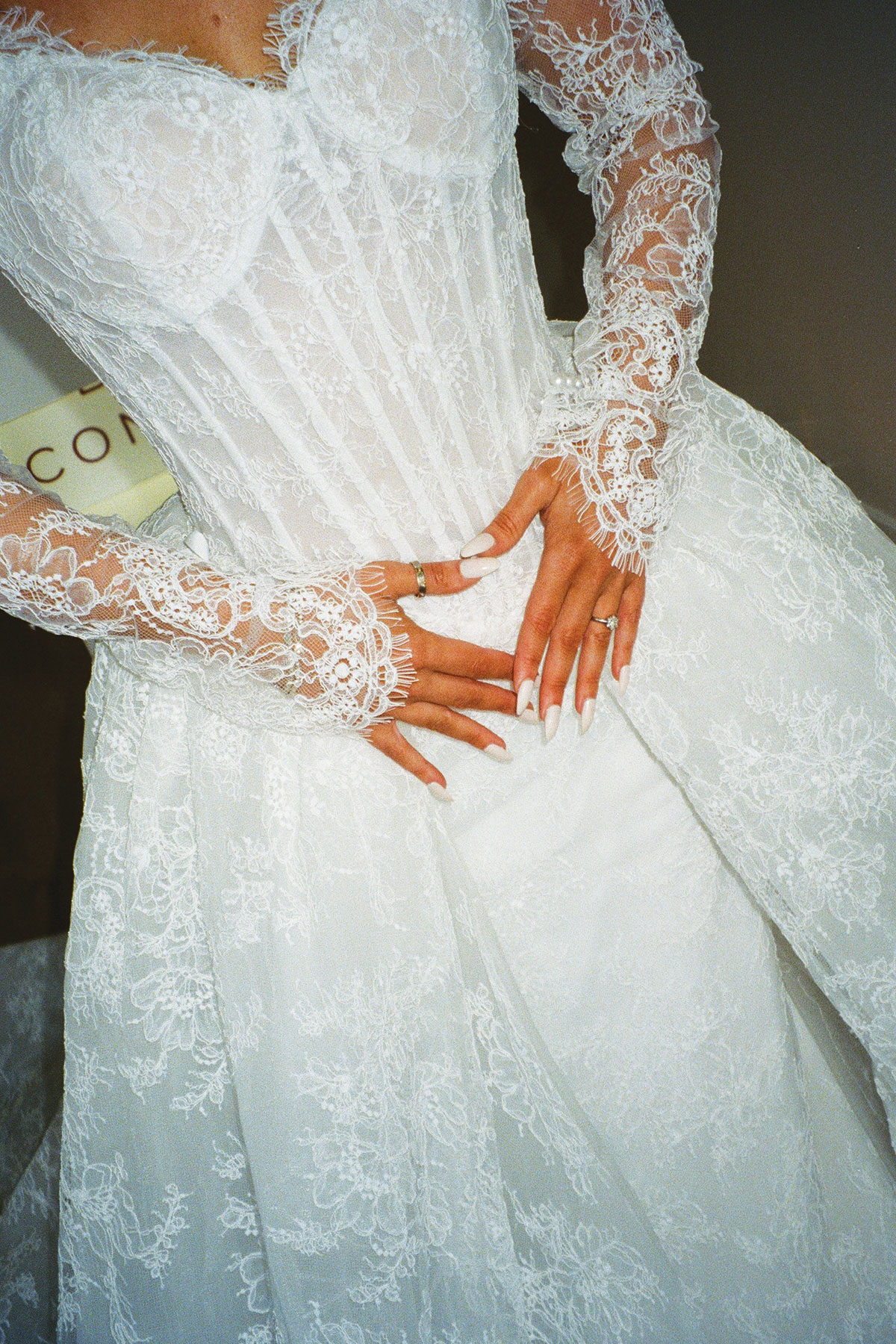 neck-down view of long-sleeved lace wedding dress with boned corset bodice and manicured nails resting at the centre of the waist
