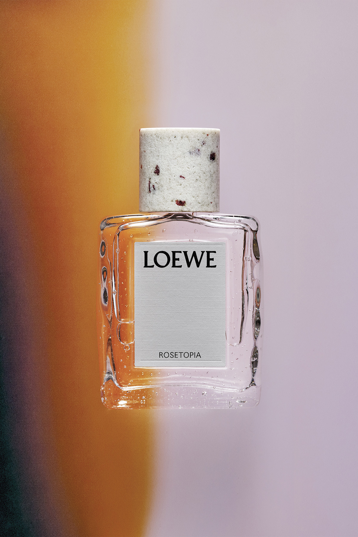 LOEWE Crafted Collection Rosetopia fragrance bottle photographed against a pink, amber and lilac background.