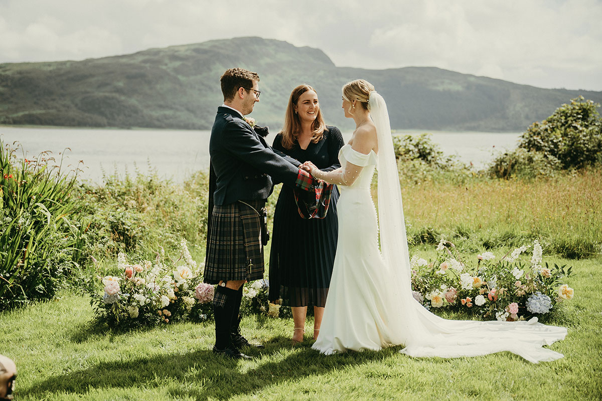 Couple hold hands during an outdoor wedding ceremony beside a loch, framed by soft pastel flowers and mountain views