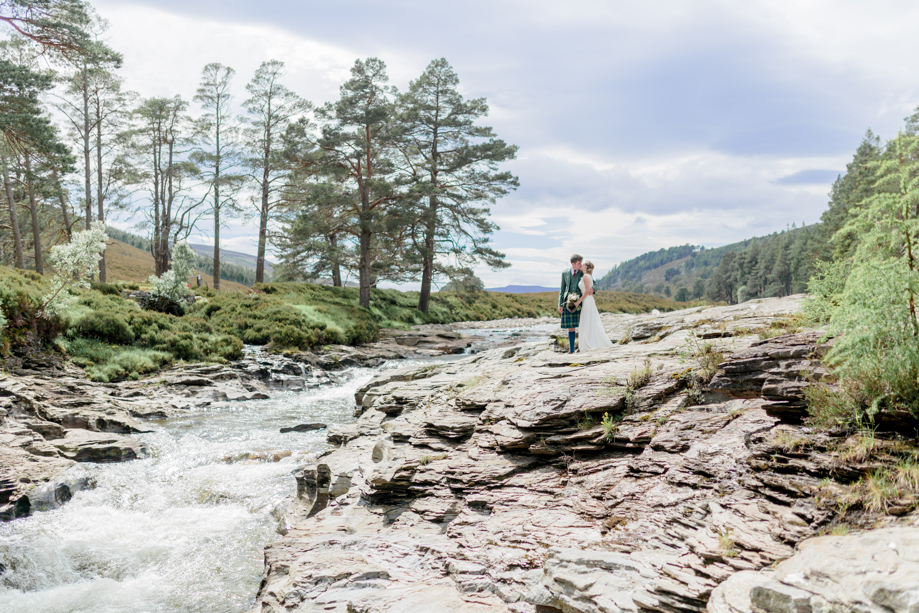 couple in wedding outfits standing on rock formation near waterfall 