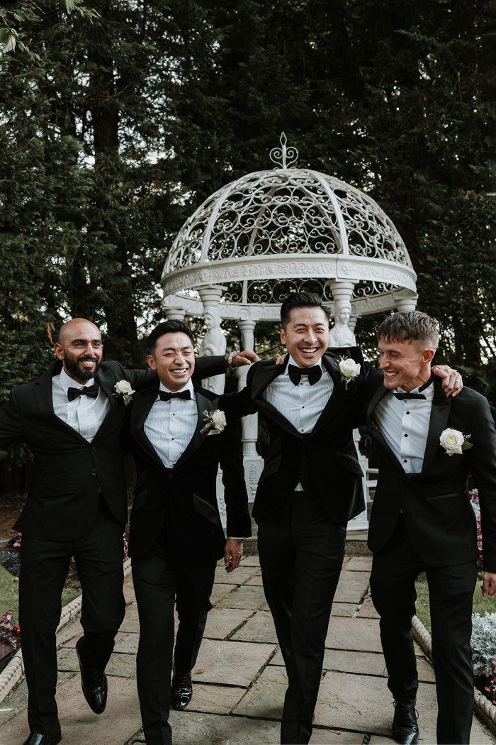 four men in black tuxedo suits walking in front of white ornamental pergola in a garden setting