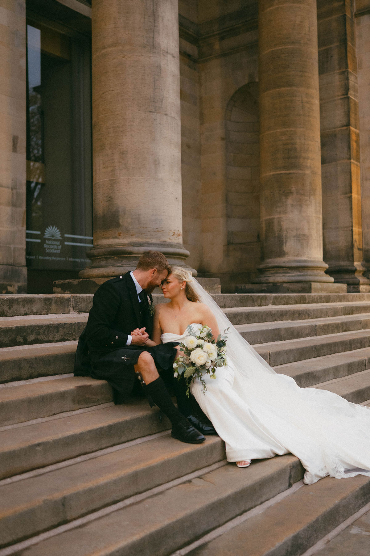 bride and groom sitting on stone steps outside historic Edinburgh venue with bouquet