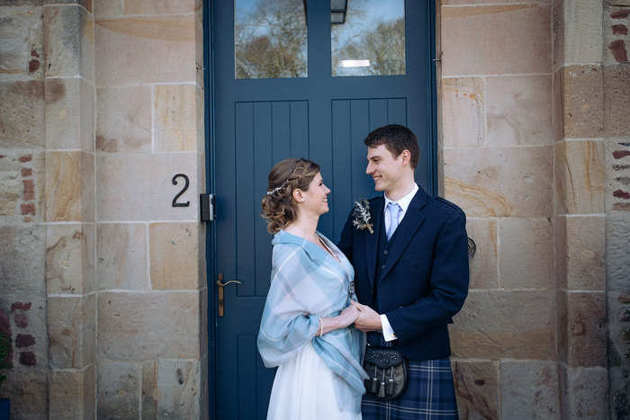 Bride wearing a tartan shawl and groom wearing kilt pose outside Newhall Mains
