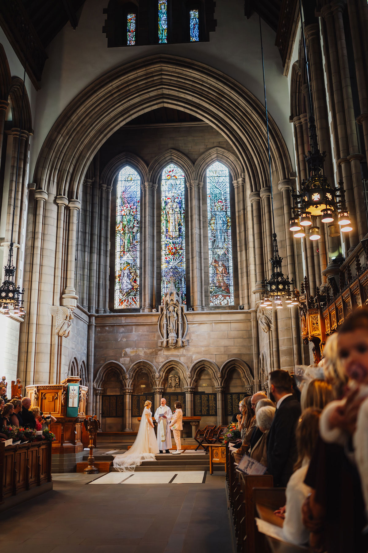 Wide view of the University of Glasgow Memorial Chapel interior showing the couple standing with the priest at the altar beneath tall arched stonework and stained-glass panels.