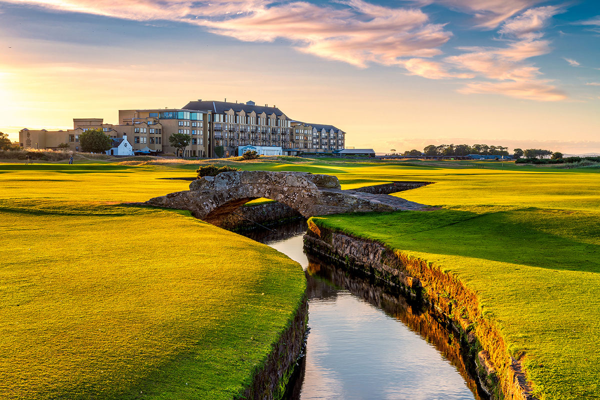 small stone bridge goes across small river from one golf grass pitch to another with a large hotel building in the background 