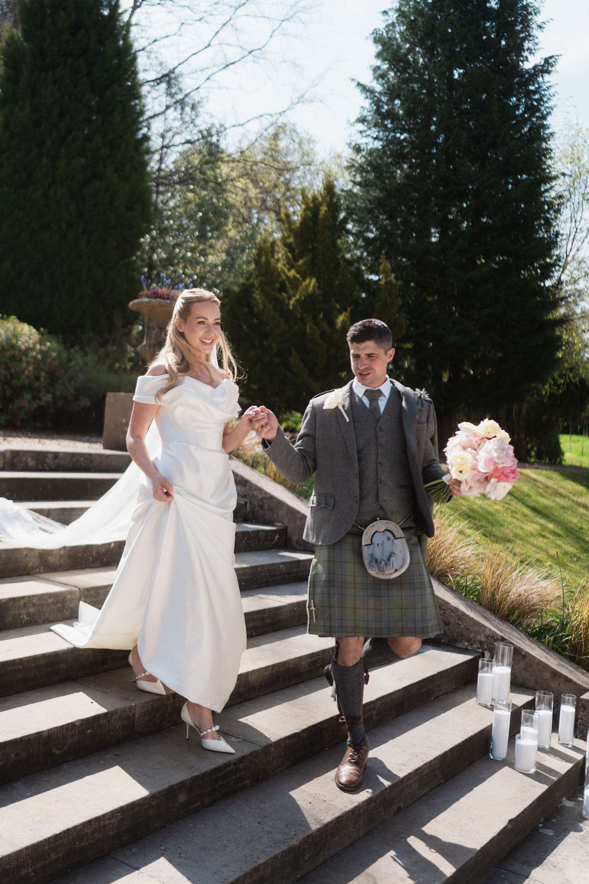 Siân and Stuart walk down the garden steps at Mar Hall, with the bride carrying a pastel pink bouquet and groom wearing a kilt.