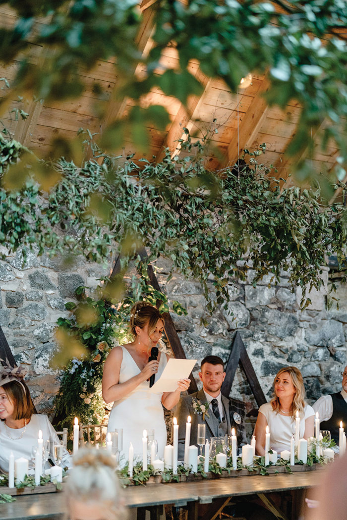 bride with her hair up and v-neck dress stands behind a long table filled with candlesticks and greenery, holding a microphone and reading from a piece of paper