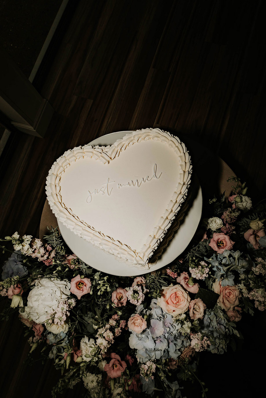 a white heart shaped cake with just married icing surrounded by pink, blue and white flowers