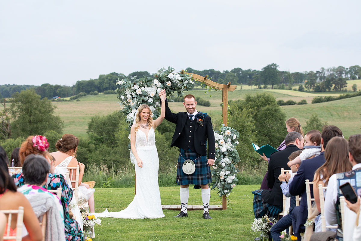Outdoor Scottish wedding ceremony with bride and groom under a floral arch in the countryside