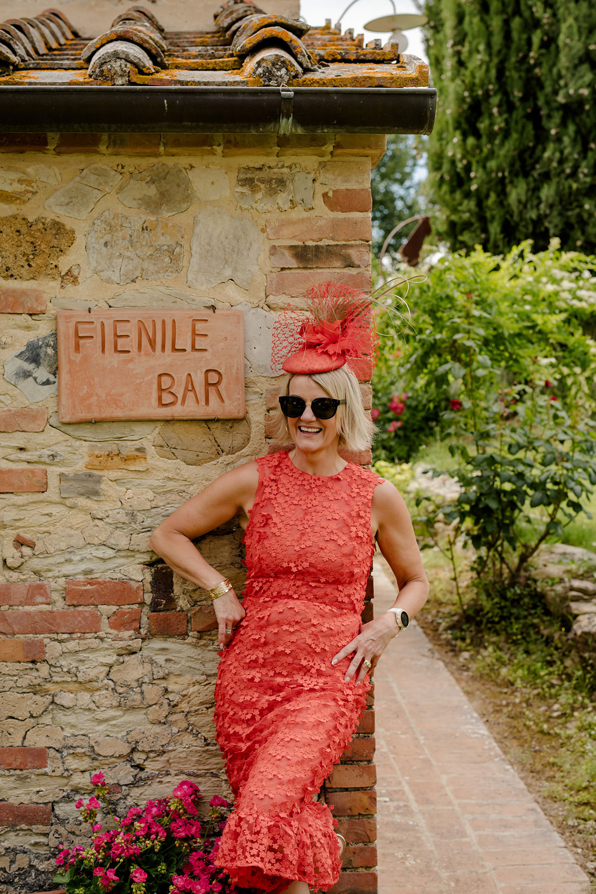 blonde woman with bob smiles and leans against brown outdoor brick wall, wearing sunglasses, a red-orange dress with 3D flowers and a matching percher hat decorated with veiling, quills and feather flowers