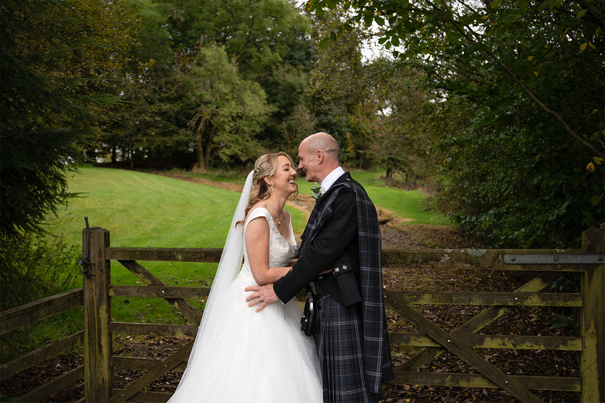 A bride and groom laughing while looking at each other and standing next to a wooden gate
