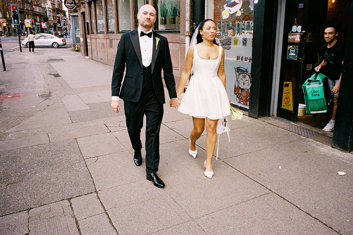 Bride and groom walking along Glasgow city street after wedding reception bride holding bouquet and groom in tuxedo