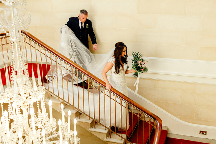 Bride walks downstairs with her dad walking behind holding veil off the ground