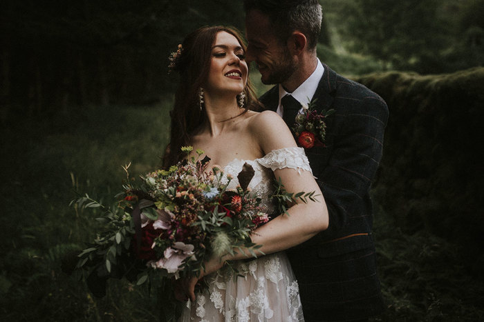 dark image of a bride and groom stood one in front of the other, the bride holding a large bouquet and leaning back over her shoulder to look at the groom