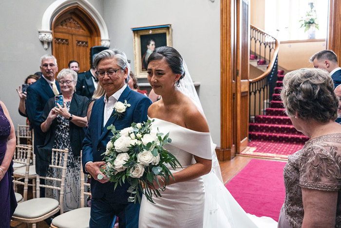 An Emotional Bride Carrying A Large White Bouquet Walks Down A Red Carpet At Blairquhan Castle On The Arm Of A Person Wearing A Navy Blue Suit As Guests Watch On