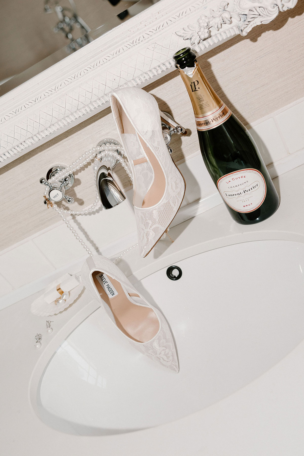 Bridal accessories including lace heels, pearl jewellery and a bottle of champagne arranged around a bathroom sink