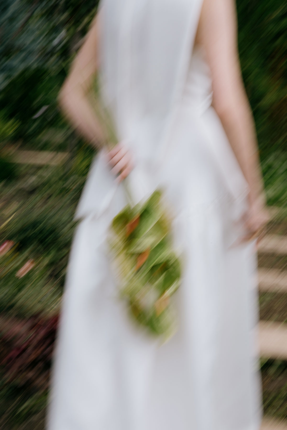 A blurred artistic shot of a bride in a white dress holding a single green anthurium stem, photographed outdoors with soft motion.