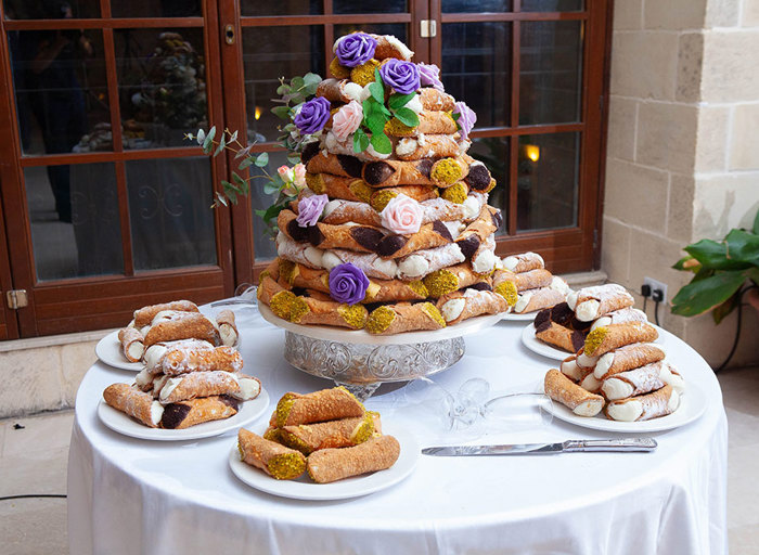 a tower of cannolis decorated with roses on a plate surrounded by smaller plates of cannoli. They sit on a round table with white tablecloth