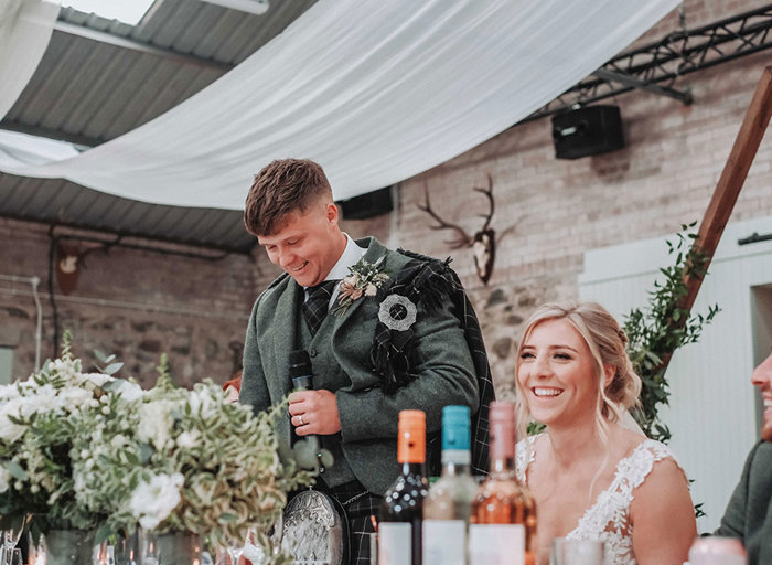 a groom standing in a stone barn setting making a wedding speech. The bride is seated at the top table next to him. There are wine bottles and green and white flowers in the foreground