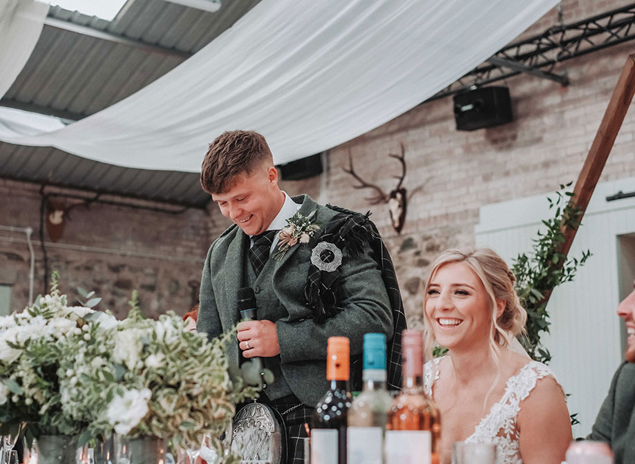 a groom standing in a stone barn setting making a wedding speech. The bride is seated at the top table next to him. There are wine bottles and green and white flowers in the foreground