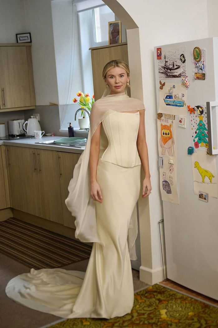 blonde woman stands in kitchen next to fridge covered in children's drawings wearing a cream wedding outfit consisting of a strapless corset bodice and silky skirt with a matching neck scarf cast over the shoulders