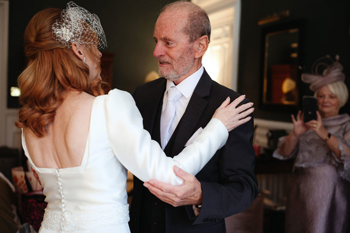 A man in a suit and a woman in a white dress look at each other with their arms around each other 