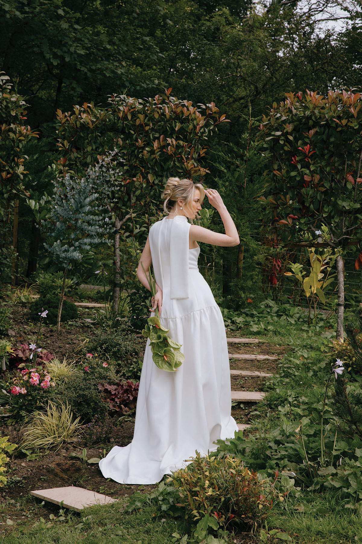 A bride in a contemporary white gown stands by a pond in a garden, holding a single anthurium flower with trees behind her.