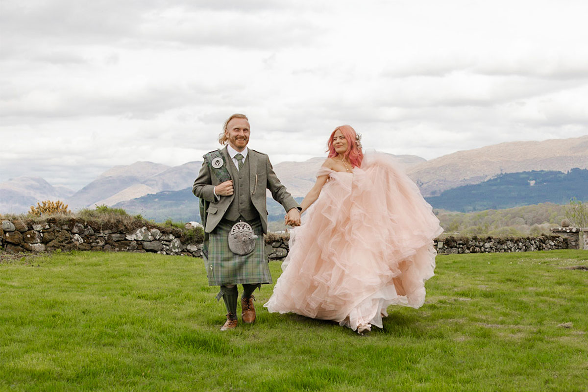 A man wearing a light green kilt and a woman wearing a pink wedding dress walking through a field with hills in the background