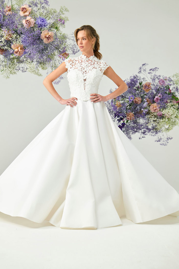 bridal model stands hands on her hips while wearing white gown with large skirt and floral detailing across the shoulders and neck