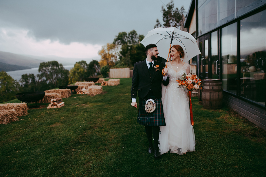 Bride and groom stroll outside under an umbrella
