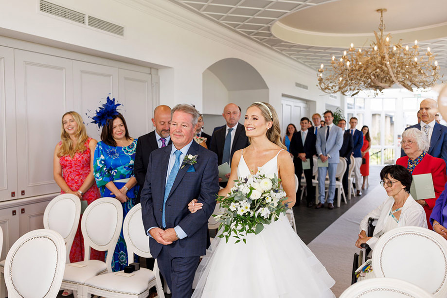 A joyful bridal procession at the Old Course Hotel: the bride, holding a bouquet of white flowers, walks down the aisle with her father, surrounded by smiling friends and family on her wedding day