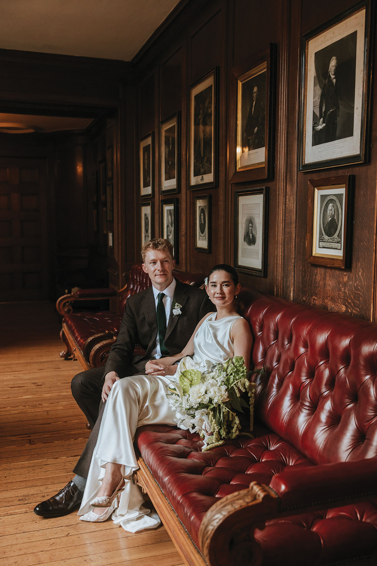 Bride and groom sitting on a red leather couch, smiling and holding bouquet.