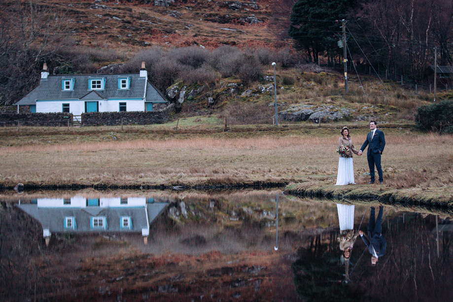 Couple hold hands beside a body of water with a house in the background 