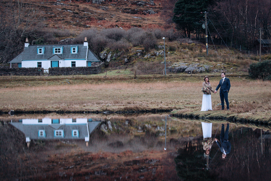 Couple hold hands beside a body of water with a house in the background