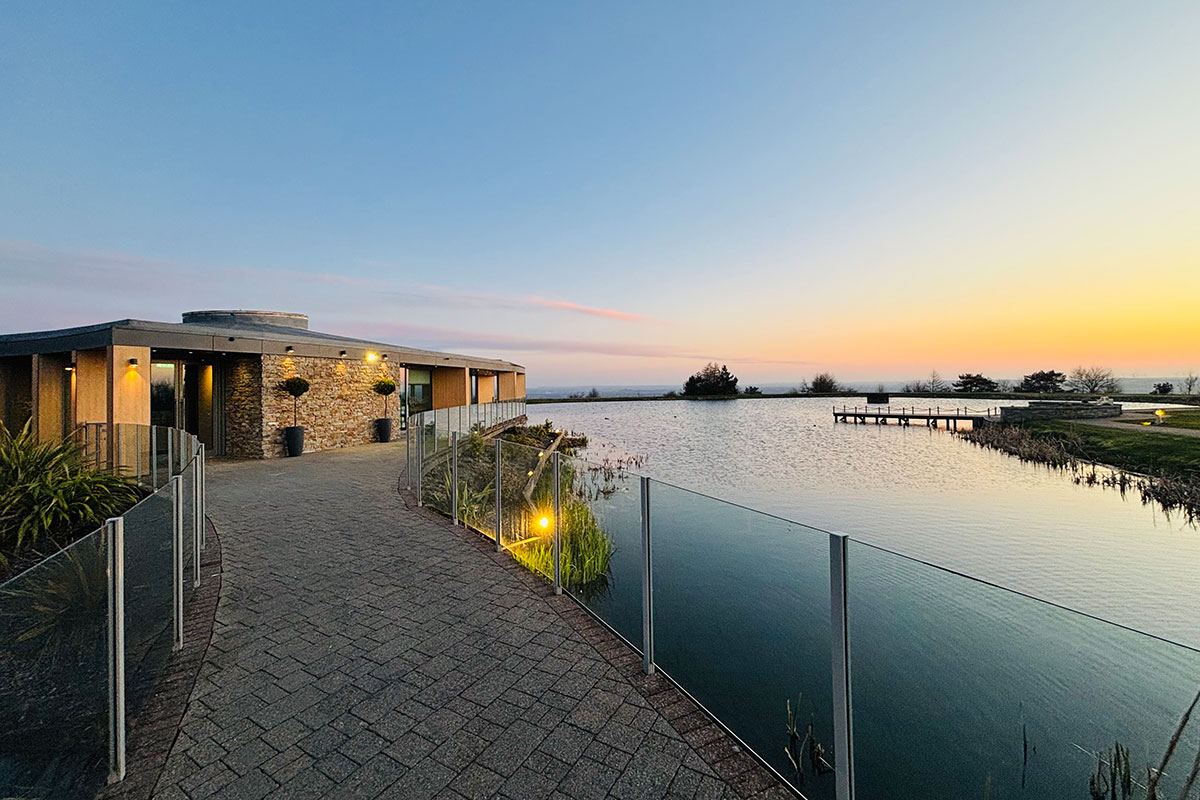 one level stone building has a grey brick walkway with glass barrier leading up to it with a view over water with a sunset coloured sky