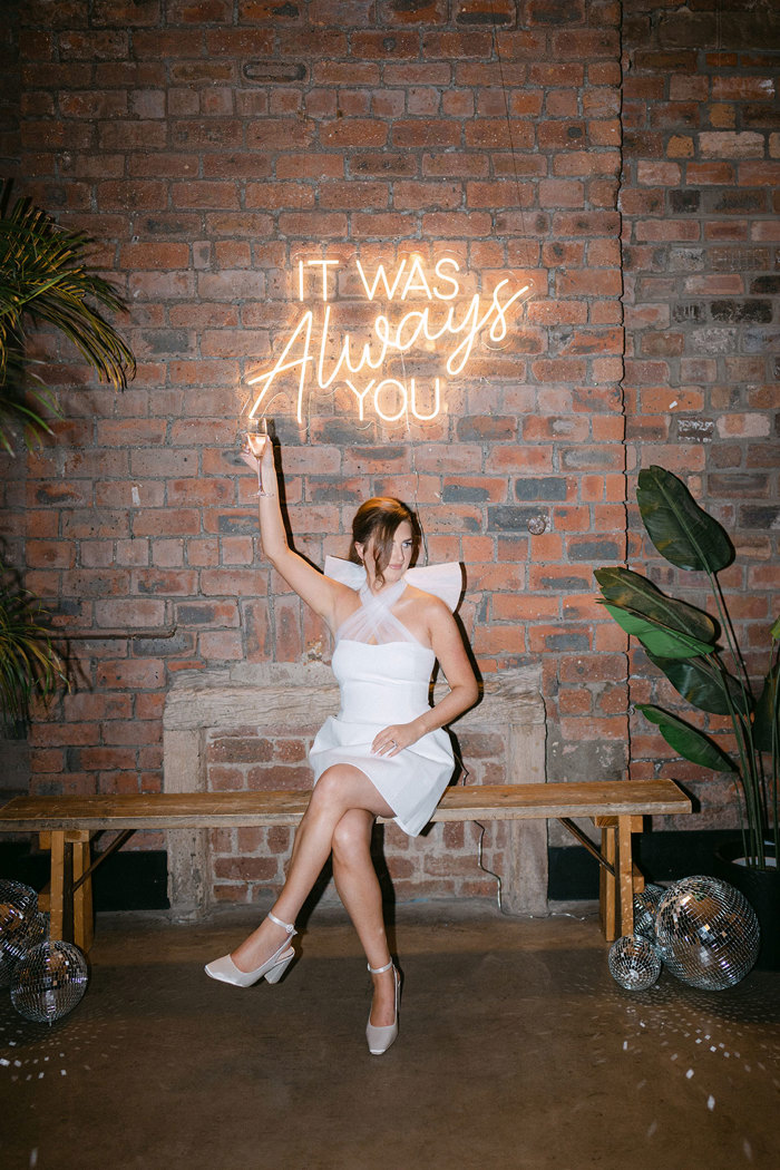 A bride sitting on a bench with a neon sign. She is wearing a short white dress with bow on the back and raising a champagne glass.