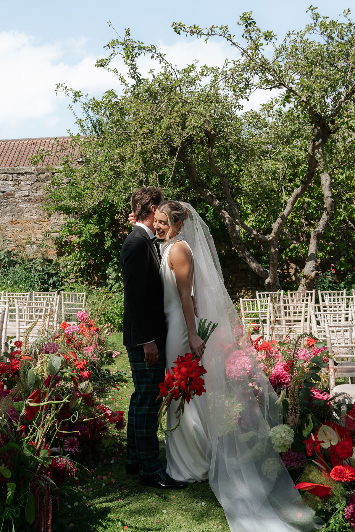 Newlyweds embrace among vibrant red and pink flowers in Kinkell Byre's orchard garden following their outdoor wedding ceremony.