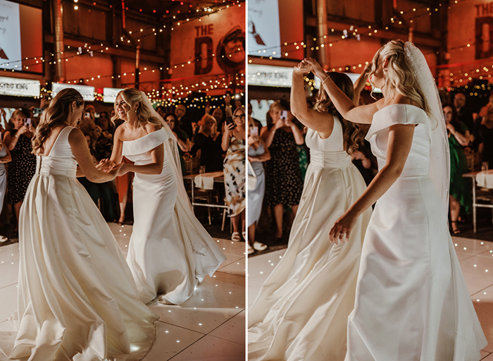 Two brides dancing on a white illuminated dance floor with fairy light canopy in the background. Guests standing in background taking pictures on smart phones.