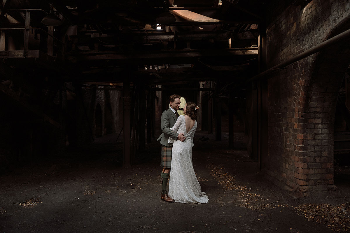 Newlyweds embrace in a dramatic industrial setting with exposed brick walls, steel structures and atmospheric lighting