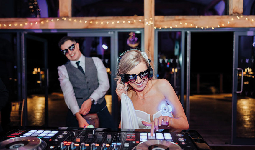 a person wearing sunglasses and headphones working at a DJ deck and a person in a suit and tie wearing sunglasses in background