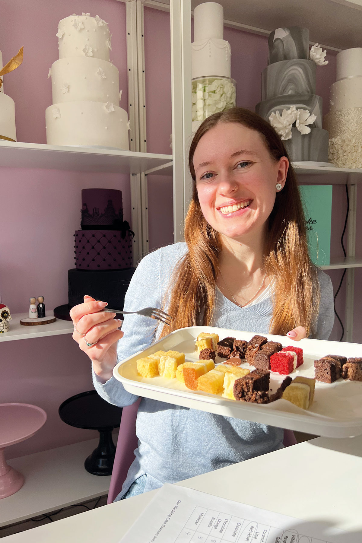 brown haired woman sits in front of open shelves of wedding cakes holding a small tray of lots of different flavours of cake samples, smiling and holding a fork with the sun on her face