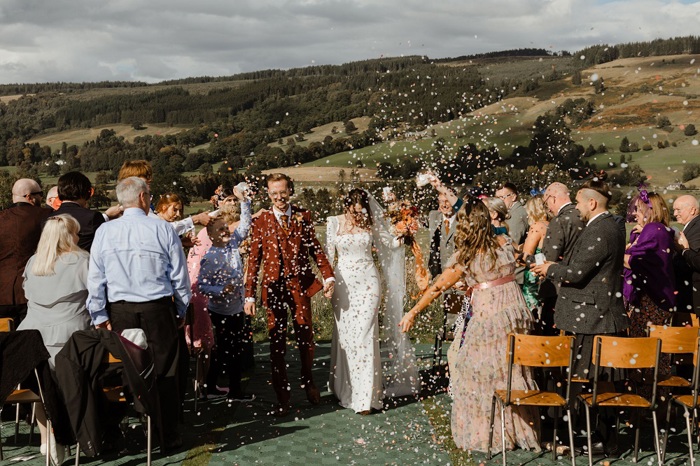 bride and groom walk down aisle of outdoor wedding as people throw confetti