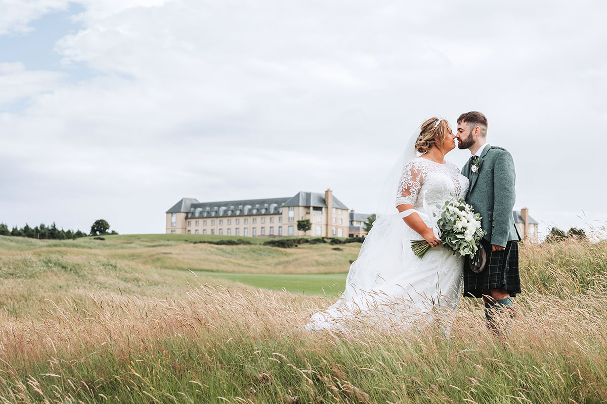Bride and groom kissing in tall grass at Fairmont St Andrews with hotel in background