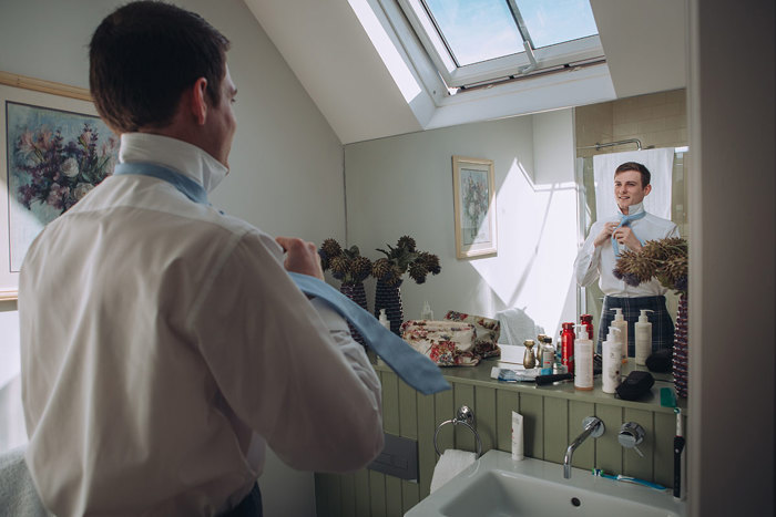 Groom tieing his tie whilst looking in the mirror