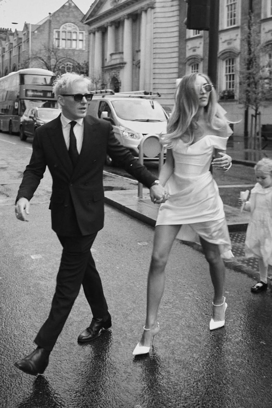 black and white image of groom in dark suit and sunglasses and bride in short off the shoulder dress and sunglasses hold hands and look to the same side as they cross a city road