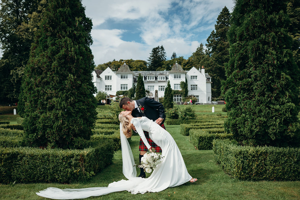 Bride and groom sharing a kiss in the formal gardens at Achnagairn Castle, with the Highland wedding venue in the background