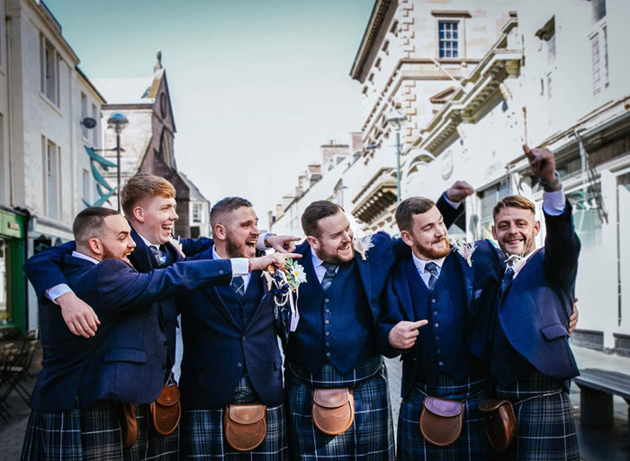 Six men in blue kilts stand with their arms around each other pointing at the man furthest on the right