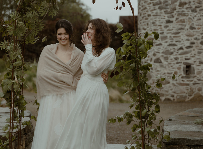 two laughing brides standing outside under a foliage-covered wire arch in a garden with old stone building in background